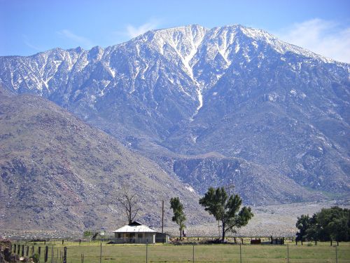 A snow-capped mountain towers over a small house and green fields under a clear blue sky.