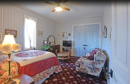 Cozy bedroom with floral decor, a bed, armchair, dresser, and a TV, featuring patterned wallpaper and a ceiling fan.