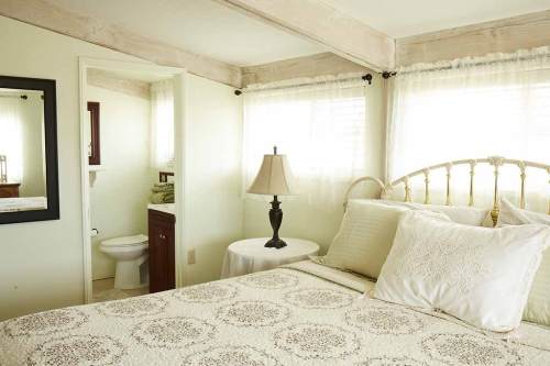 Cozy bedroom with a bed, lamp, and a bathroom visible in the background, featuring soft natural light.