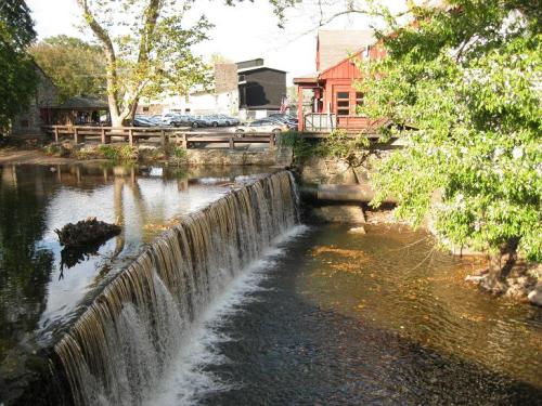 A serene waterfall flows into a calm river, with a red building and trees in the background.