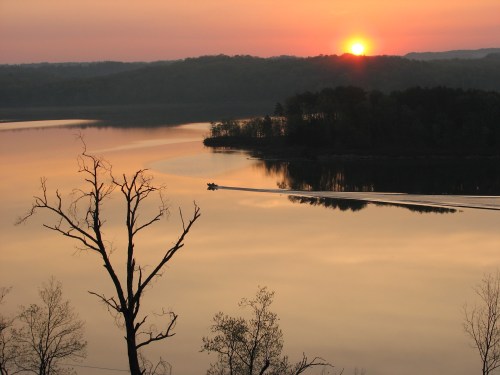 A serene sunset over a calm lake, with silhouettes of trees and a small boat gliding through the water.