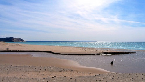 A serene beach scene with gentle waves, soft sand, and a clear blue sky reflecting sunlight over the water.