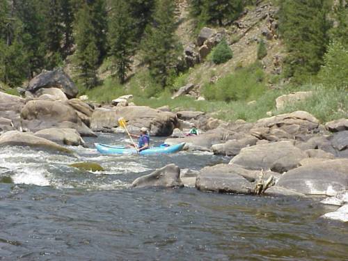A person kayaking through rocky rapids surrounded by trees and greenery.