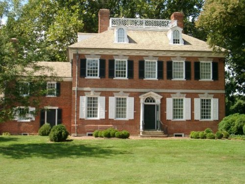 A large, historic brick house with white shutters, a central door, and a decorative roof, surrounded by green lawns.