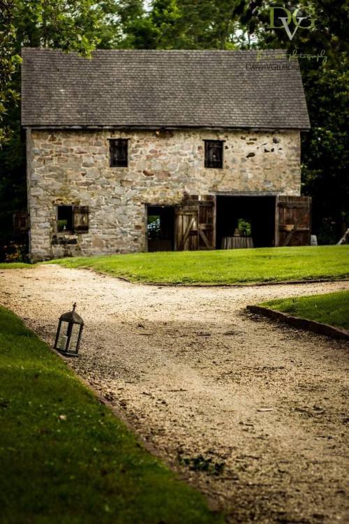 A rustic stone building with a sloped roof, surrounded by greenery and a gravel path leading to it.
