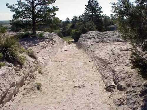 A sandy pathway cuts through rocky terrain, flanked by sparse vegetation and trees under a clear blue sky.