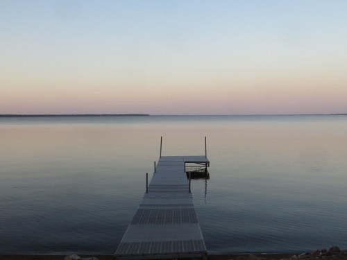 A calm lake at dusk with a wooden dock extending into the water, reflecting soft pastel colors in the sky.