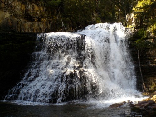 A cascading waterfall flows over rocky tiers, surrounded by lush greenery and steep cliffs.