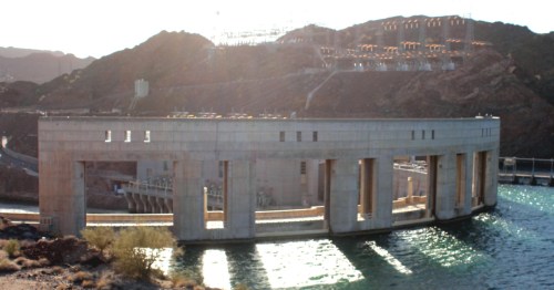 A large concrete structure on the water's edge, surrounded by rocky hills and power lines in the background.