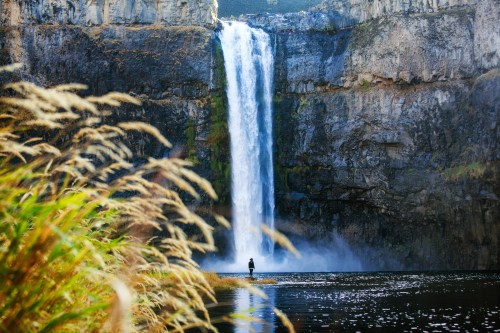 A person stands near a tall waterfall surrounded by rocky cliffs and lush greenery, with mist rising from the water.