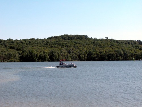 A small boat glides across a calm lake, surrounded by lush green hills under a clear blue sky.