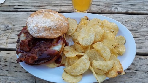 A plate with a sandwich topped with bacon and cheese, alongside a serving of crispy potato chips and a drink.