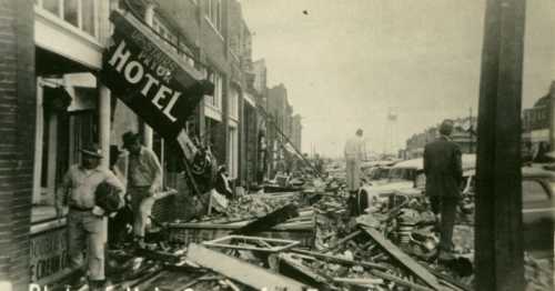 Historic photo of a street after a disaster, featuring debris, damaged buildings, and a fallen hotel sign.