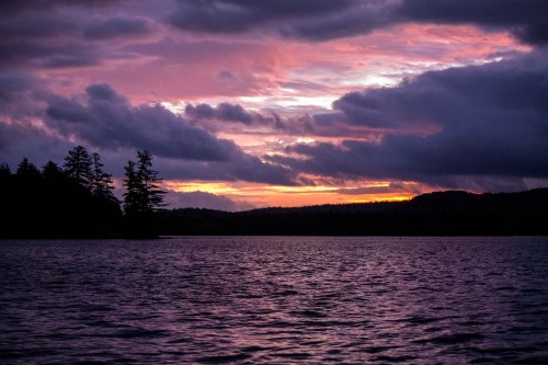 A serene lake at sunset, with vibrant purple and orange skies, silhouetted trees, and gentle ripples on the water.