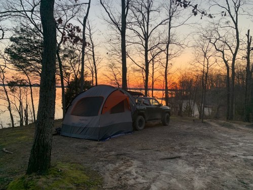 A tent set up by a lake at sunset, surrounded by trees and a rugged vehicle nearby.