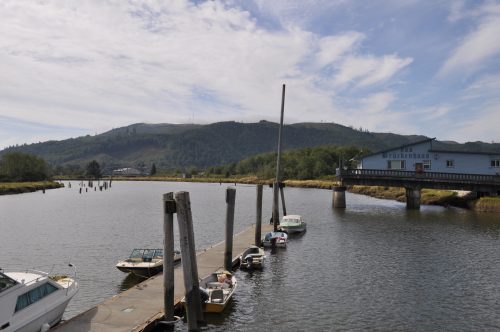 A calm waterway with boats docked along a pier, surrounded by green hills and a building in the background.