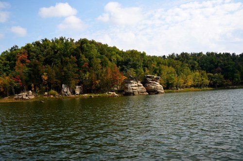 Scenic view of a calm lake surrounded by colorful autumn trees and rocky formations under a partly cloudy sky.