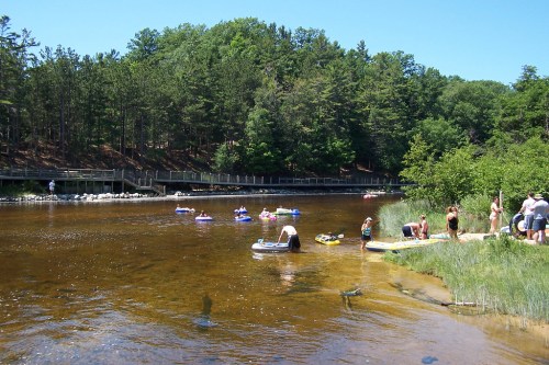 People enjoying a sunny day by a river, with some swimming and others relaxing on the shore surrounded by trees.