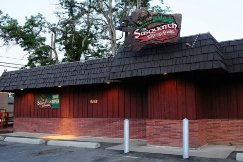 Exterior of The Sasquatch Tavern & Grill, featuring a wooden facade and a sign, with trees in the background.