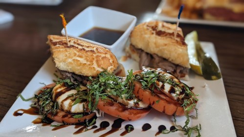 A plate featuring two sandwiches with toothpicks, a dipping sauce, and sliced tomatoes topped with herbs and balsamic drizzle.