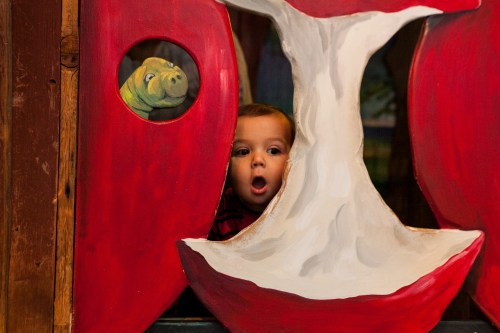 A baby peeks through a painted apple cutout, with a cartoon character visible in the background.