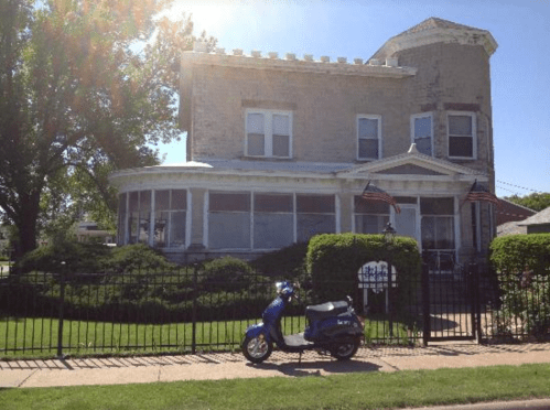 A large, historic house with a wraparound porch, surrounded by greenery, and a blue scooter parked in front.
