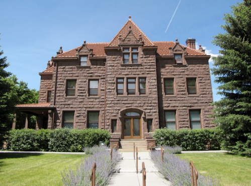 A large, historic brownstone building with a red tile roof, surrounded by greenery and a clear blue sky.