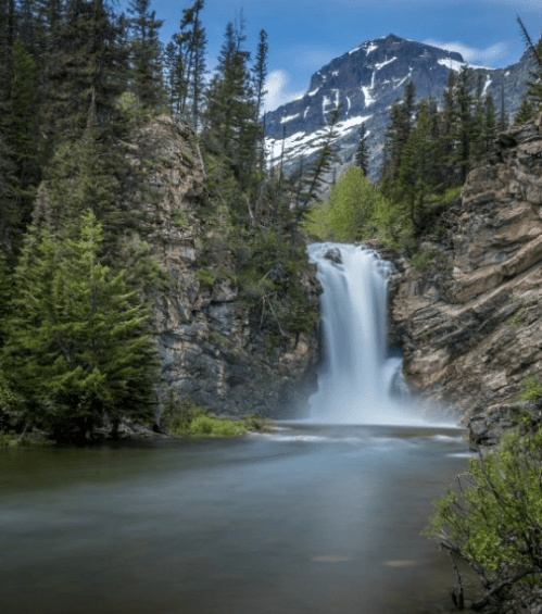 A serene waterfall cascades down rocky cliffs, surrounded by lush greenery and towering mountains under a clear sky.