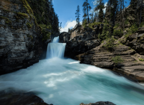 A serene waterfall cascades through rocky cliffs, surrounded by lush green trees under a clear blue sky.