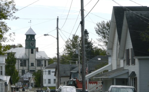 A small town street with power lines, houses, and a clock tower in the background under a cloudy sky.