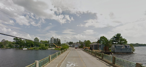 A scenic view of a bridge over water, with trees and buildings lining the shore under a cloudy sky.