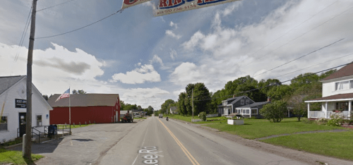 A quiet rural road with a banner overhead, flanked by buildings and trees under a partly cloudy sky.
