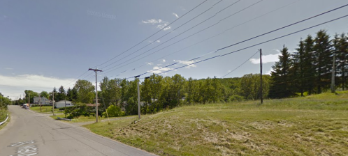 A rural road with power lines, grassy areas, and trees in the background under a clear blue sky.