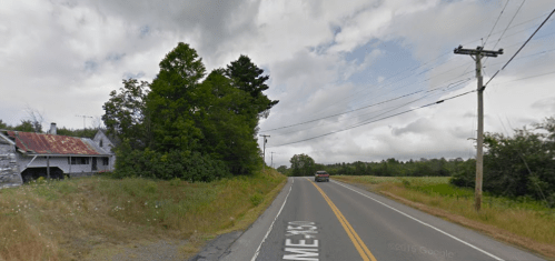 A rural road with a weathered house and trees on one side, under a cloudy sky.