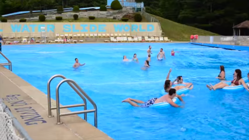 A lively water park scene with people swimming and floating in a blue pool, surrounded by slides and lounge chairs.