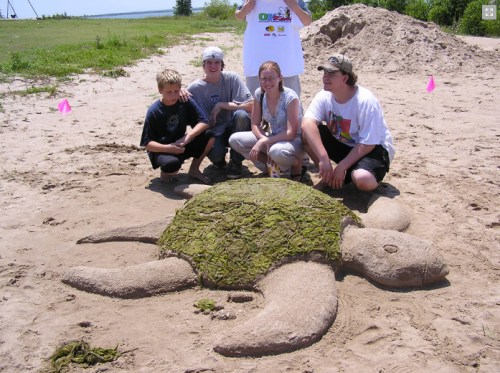 A group of four people poses beside a large sand sculpture of a turtle, decorated with green seaweed, on a beach.
