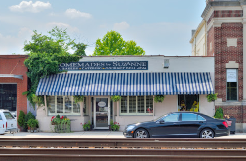 A charming bakery and catering shop with a blue-striped awning, surrounded by greenery and parked cars nearby.