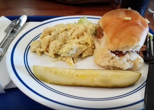 A plate with a sandwich, pasta salad, and a pickle slice, served on a blue tray with utensils.