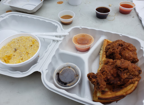 A takeout container with fried chicken on a waffle, grits, and several small sauce cups on a table.