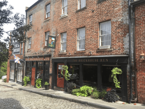 Historic brick buildings with flower planters, featuring "The Brickhouse Run" sign and a British flag. Cobblestone street.