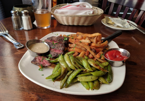 A plate with sliced steak, green beans, and fries, served with dipping sauce and ketchup, on a wooden table.