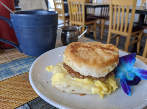 A heart-shaped biscuit sandwich with sausage and scrambled eggs, served with a blue flower and a cup of coffee.