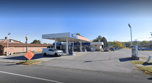 A gas station with a convenience store, featuring pumps and a sign, set in a suburban area with clear skies.