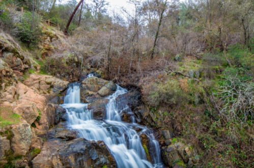 A serene waterfall cascades over rocky terrain, surrounded by trees and lush greenery in a tranquil natural setting.