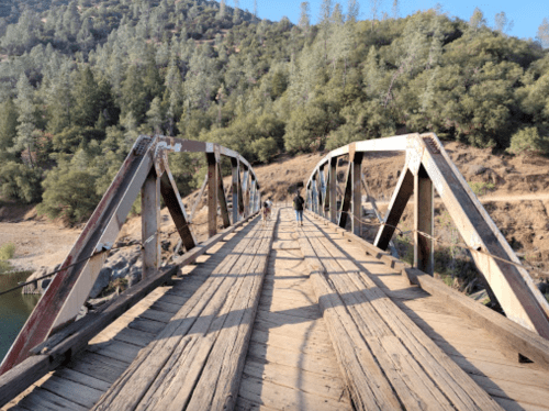 A wooden bridge spans a river, with two people walking towards the camera amidst a forested landscape.