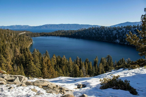A serene lake surrounded by snow-covered trees and mountains under a clear blue sky.