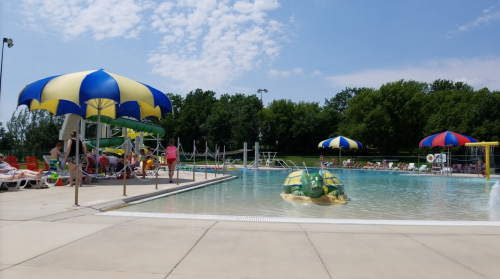 A sunny pool scene with colorful umbrellas, lounge chairs, and people enjoying the water and surrounding area.