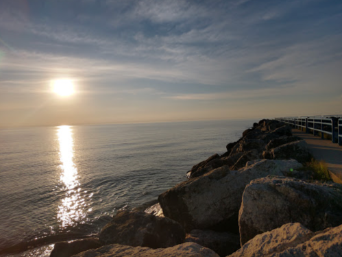 A serene view of the sun setting over calm waters, with a rocky shoreline and a railing in the foreground.