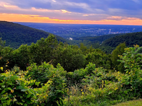 A scenic view of rolling hills and a valley at sunset, with vibrant colors in the sky and lush greenery in the foreground.