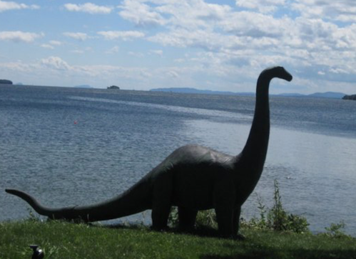 A dinosaur statue stands by a lake under a cloudy sky, with mountains visible in the distance.